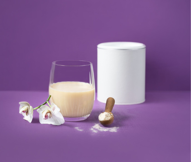 Wasserglas gefüllt mit Vanillemilch, davor ein Holzlöffel mit Pulver und eine Orchidee