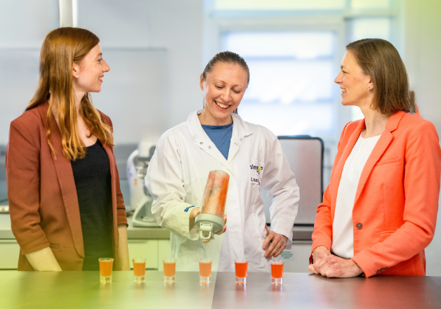 3 Kolleginnen unterhalten sich am Tisch während eine von Ihnen einen Proteinshake zubereitet